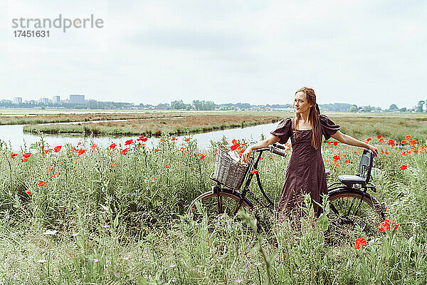 Frau mit Fahrrad steht zwischen Mohnblumenfeld vor dem Himmel
