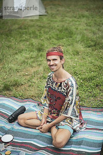 Lächelnder Mann im seidenen Drachenoberteil sitzt auf einer Picknickdecke im Camp im Gras