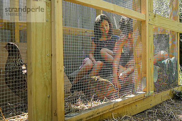 Zwei zufriedene Kinder sitzen zusammen im Hühnerstall und spielen mit Hühnern