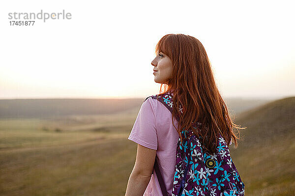 Junge rothaarige Frau mit Rucksack schaut im Sonnenuntergang weg