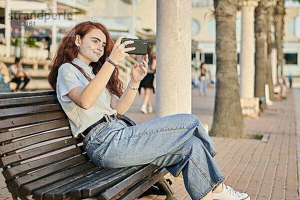Junge rothaarige Frau macht ein Foto mit ihrem Handy  das auf einer Bank sitzt