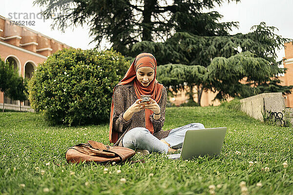 Muslimische Frau sitzt mit Computer und Telefon im Gras
