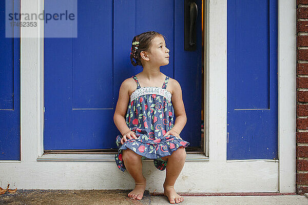 Süßes kleines Mädchen mit Zöpfen sitzt barfuß im Sommerkleid auf der Vordertreppe