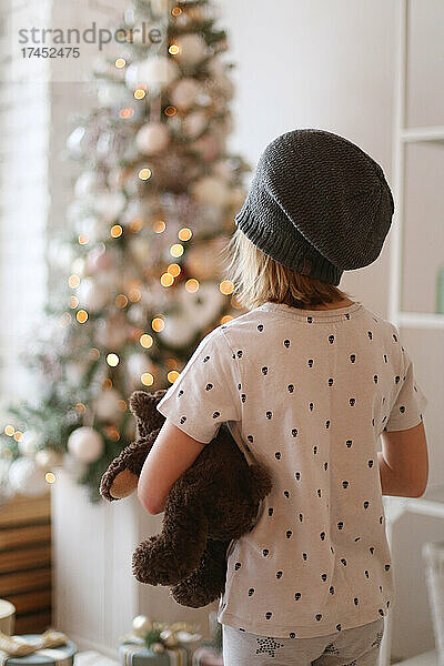 Ein Kind mit Hut steht vor einem Weihnachtsbaum