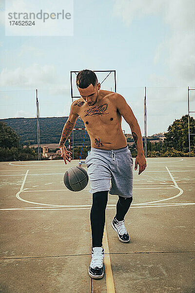 Junger tätowierter Latino-Junge spielt mit einem Basketball auf einem Platz