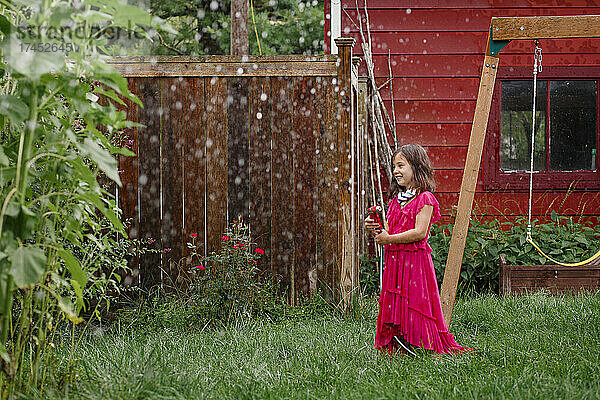 Ein kleines Mädchen in einem langen bunten Kleid spielt im Hinterhof mit einem Schlauch