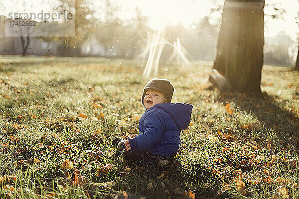 Kleiner Säugling in blauer Jacke und warmer Mütze sitzt auf dem Gras