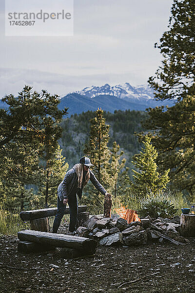Frau in Puffjacke legt Holzscheite auf Lagerfeuer in den Bergen
