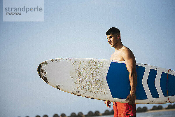 Junger Mann hält sein Surfbrett am Strand.