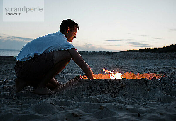 Mann entzündet fröhlich ein Lagerfeuer am Strand bei Sonnenuntergang