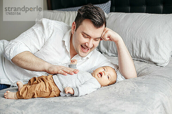 Glücklicher stolzer Vater  der auf dem Bett liegt und mit seinem neugeborenen Jungen spielt