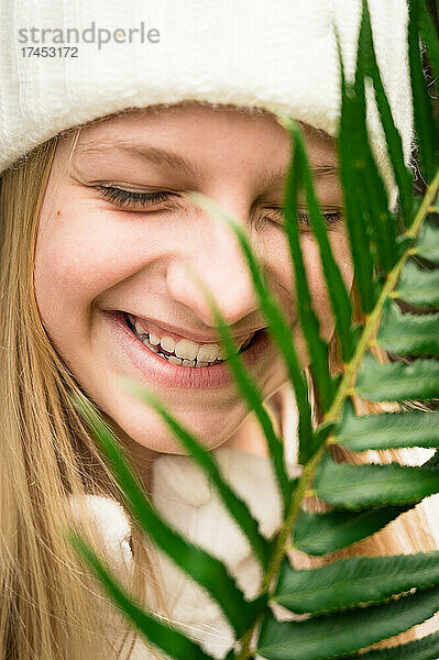 Junges Mädchen mit Wintermütze lacht hinter grünem Farn