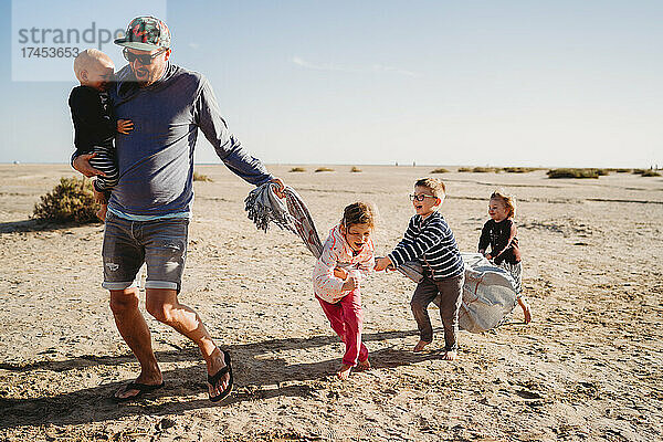 Vater und glückliche Kinder spielen an einem kühlen Tag mit einem Handtuch am Strand