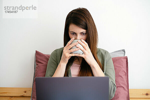 Junge Frau schaut sich Videos per Laptop an und trinkt Kaffee.