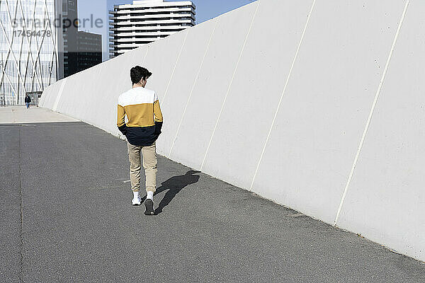 Rückansicht eines jungen Mannes  der neben einer Wand geht und die Hände auf die Tasche legt