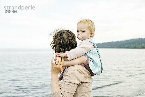 Ausgeschnittene Ansicht eines Kleinkindes  das auf den Schultern seiner Mutter am Strand sitzt