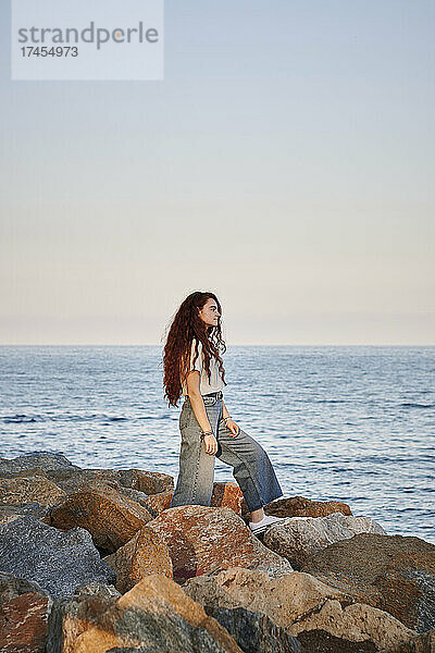 Junge rothaarige Frau blickt auf das Meer  während sie auf Felsen steht