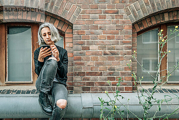 Eine Frau sitzt in der Nähe eines alten Backsteingebäudes und benutzt ein Smartphone