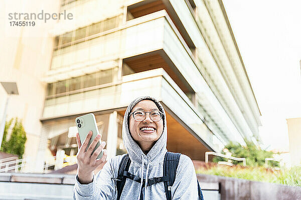 Junge asiatische Person hält Smartphone lächelnd in der Innenstadt