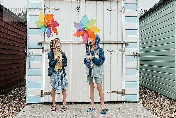 Geschwister lachen und spielen mit Windmühlen neben Strandhütten