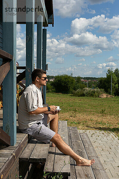 Ein junger Mann sitzt mit einer Tasse Kaffee auf der Veranda eines Hauses