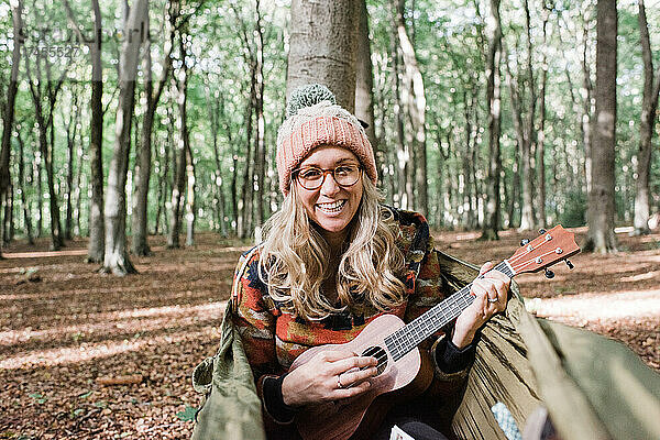 Frau lacht  während sie auf einer Hängematte im Wald Ukulele spielt