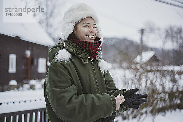 Lächelnde nicht-binäre Person zieht im schneebedeckten Dorf Winterhandschuhe an