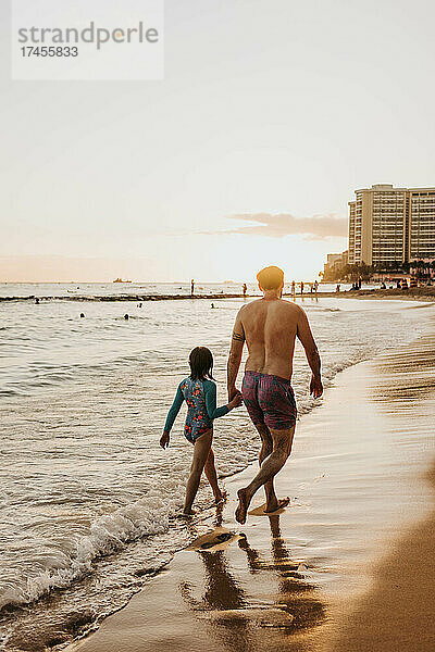 Vater und Tochter halten Händchen  während sie bei Sonnenuntergang am Strand entlang spazieren