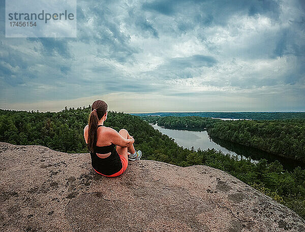 Frau sitzt auf dem Gipfel einer Wanderung mit Blick auf See und Bäume.