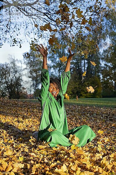Porträt einer lachended  dunkelhäutigen Frau mit Locken im grünen Kleid  sitzt und wirft Herbstlaub  Außenaufnahme im Herbst  Deutschland  Europa