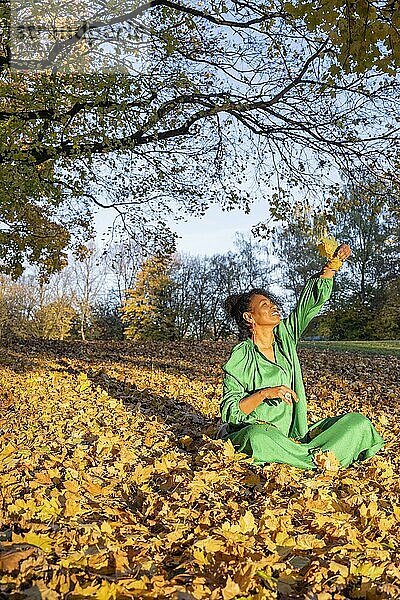 Porträt einer lächelnden  dunkelhäutigen Frau mit Locken im grünen Kleid  sitzt im Herbstlaub  Außenaufnahme im Herbst  Deutschland  Europa