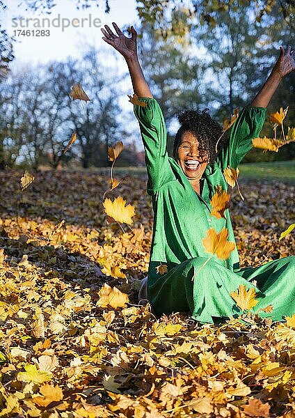 Porträt einer lachenden  dunkelhäutigen Frau mit Locken im grünen Kleid  sitzt und wirft Herbstlaub  Außenaufnahme im Herbst  Deutschland  Europa