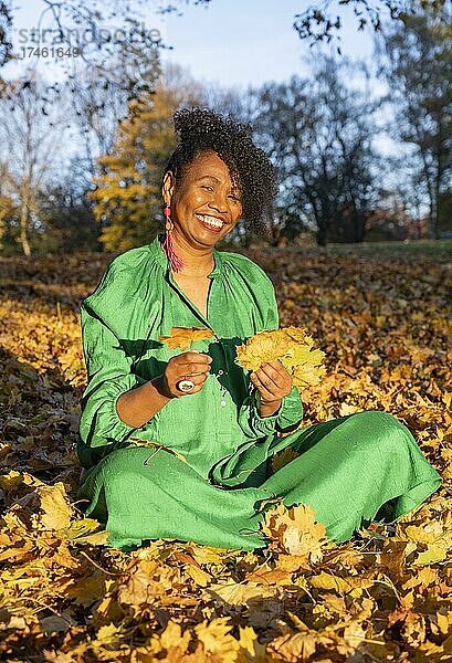 Porträt einer lächelnden  dunkelhäutigen Frau mit Locken im grünen Kleid  sitzt im Herbstlaub  Außenaufnahme im Herbst  Deutschland  Europa