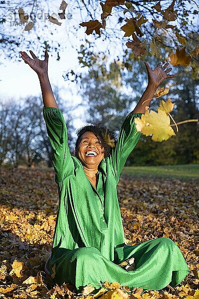 Porträt einer lachenden  dunkelhäutigen Frau mit Locken im grünen Kleid  sitzt und wirft Herbstlaub  Außenaufnahme im Herbst  Deutschland  Europa