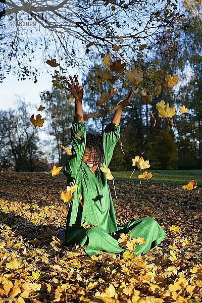 Porträt einer lächelnden  dunkelhäutigen Frau mit Locken im grünen Kleid  sitzt und wirft Herbstlaub  Außenaufnahme im Herbst  Deutschland  Europa
