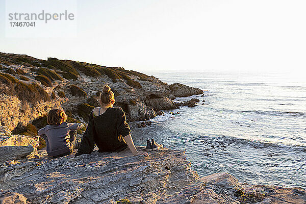 Teenager-Mädchen und junger Bruder bei Sonnenuntergang  nebeneinander sitzend mit Blick auf den Ozean.