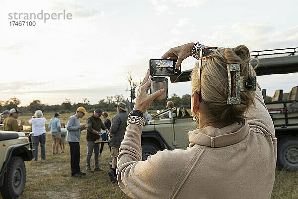 Rückansicht einer Frau  die mit ihrem Smartphone ein Foto von einer Gruppe von Menschen auf einer frühmorgendlichen Pirschfahrt macht