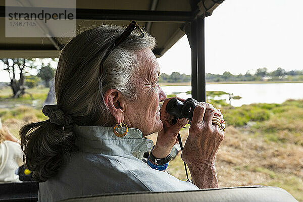 Ältere Frau mit Fernglas  in einem Safarifahrzeug sitzend  mit Blick über Sümpfe und Wasserwege