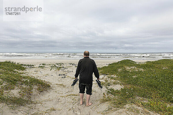 Ein reifer Mann  der mit seinen Schuhen in der Hand über einen Strand läuft