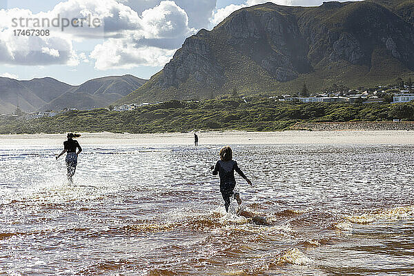 Teenager-Mädchen und ein Junge laufen durch flaches Wasser an einem breiten Sandstrand