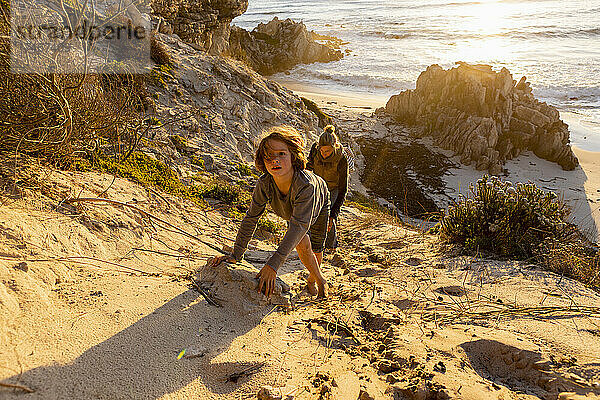 Ein Junge und eine Frau klettern einen sehr steilen Sandhang oberhalb eines Strandes hinauf