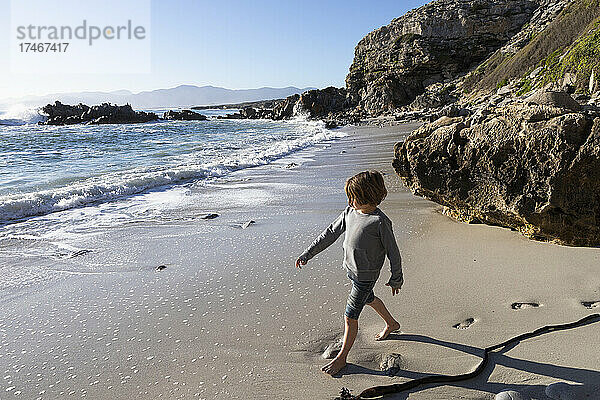 Ein kleiner Junge allein auf einem kleinen Stück Sand unter den Klippen am Meer.