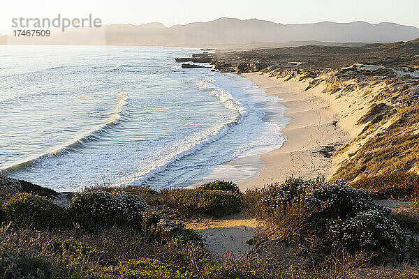 Blick von den Klippen auf den Sandstrand und die sich am Ufer brechenden Wellen.