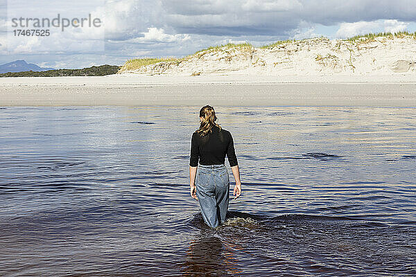 Ein junges Mädchen watet durch einen Wasserkanal an einem breiten Sandstrand.