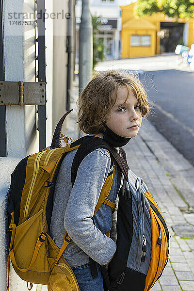 Ein Junge mit einer schwarzen Gesichtsmaske unter dem Kinn  auf der Straße mit einem Rucksack und einer Tasche.