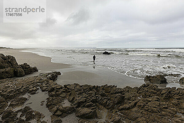 Ein Mann geht am Strand über den Sand zum Wasser  es ist bewölkt und die Wellen brechen am Ufer.