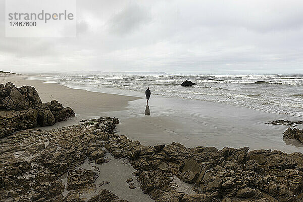 Ein Mann geht am Strand über den Sand zum Wasser  es ist bewölkt und die Wellen brechen am Ufer.