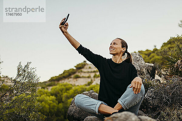 Lächelnde Frau macht ein Selfie mit ihrem Mobiltelefon auf einem Felsen