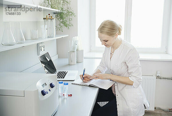 Blonder Wissenschaftler schreibt mit Laptop in Notizblock am Schreibtisch