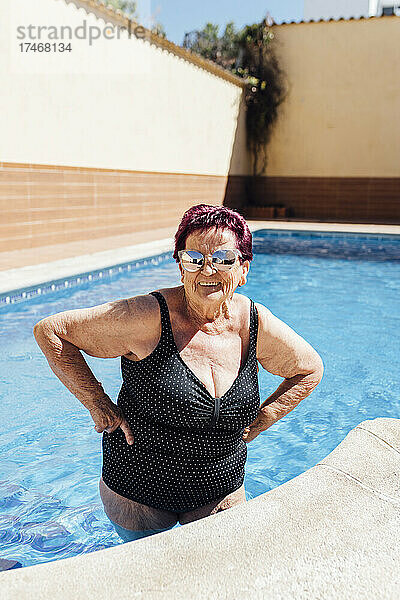 Lächelnde ältere Frau mit der Hand auf der Hüfte  die im Urlaub im Schwimmbad steht
